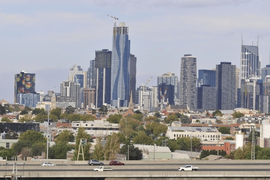 Photograph of a city skyline featuring a mix of modern high-rise buildings and mid-rise structures under a partly cloudy sky. A highway with moving cars is visible in the foreground, highlighting urban development and transportation infrastructure.