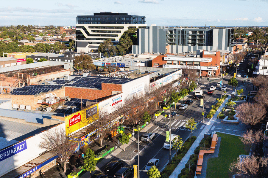 Photograph of a busy urban street in Sunshine Precinct featuring retail shops, parked cars, and pedestrian walkways. The scene includes a supermarket with solar panels on its roof, modern office buildings in the background, and landscaped green spaces with benches along the sidewalk.