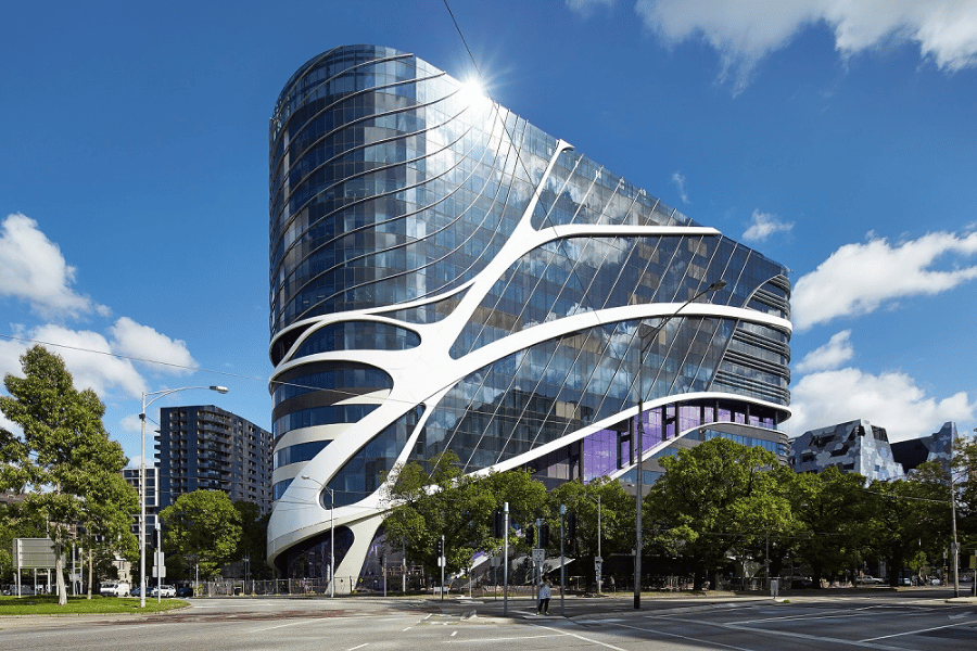 Photograph of a modern, futuristic building with curved glass facade and white structural accents resembling organic shapes. The building is surrounded by trees and urban infrastructure under a partly cloudy blue sky, highlighting innovative architectural design.