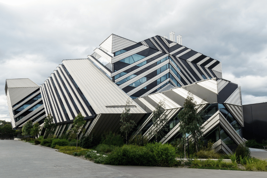 Photograph of a modern, geometric building with angular shapes and black-and-white striped patterns on its exterior. The structure features multiple levels with large windows and is surrounded by greenery under a cloudy sky.