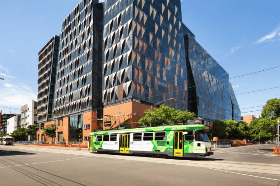 Photograph of a modern city street featuring a green and white tram traveling on tracks in front of a large, angular building with a geometric glass facade and brick base.