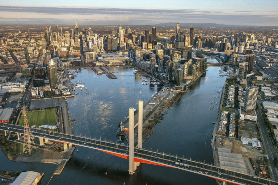 Aerial photograph showcasing a city skyline with a prominent river running through it, featuring a large cable-stayed bridge with red and white towers in the foreground. The scene highlights dense urban development with numerous high-rise buildings, waterfront docks, and a clear sky with layered clouds above.