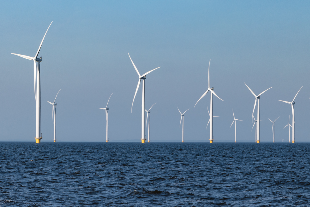 Photograph of an offshore wind farm featuring multiple white wind turbines with yellow bases standing in ocean water under a clear blue sky. The turbines are evenly spaced, highlighting renewable energy generation and sustainable power infrastructure.